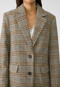 Blazer à carreaux dans des tons beige et marron, avec un col à revers cranté, deux poches avant et deux boutons noirs, confectionné dans un tissu texturé.