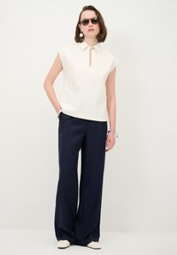 Cremfarbene ärmellose Bluse mit Schlüssellochdetail und spitzem Kragen, kombiniert mit navyblauen Weitentrosers. Das Modell trägt dunkle Sonnenbrille und Accessoires.