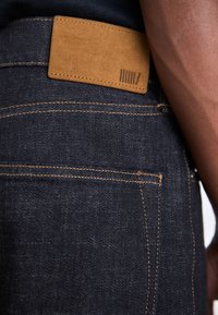 Los jeans de mezclilla oscura presentan costuras en contraste en color marrón claro y un parche de ante en la cinturilla con líneas verticales. La vista en primer plano enfatiza la textura y el ajuste.