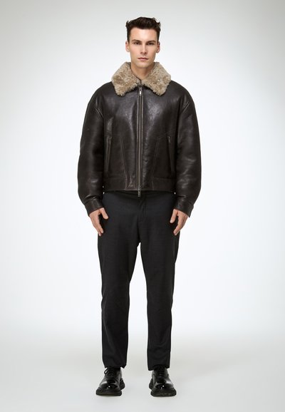 Brown leather bomber jacket with a beige shearling collar, zip closure, and side pockets, paired with dark tailored trousers and black shoes.