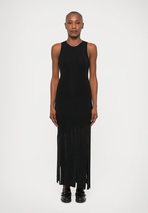 Zwarte mouwloze jurk met een geribbelde top en een asymmetrische franjeszoom. Gladde textuur, enkel-lang, aangevuld met zwarte schoenen met gespen.