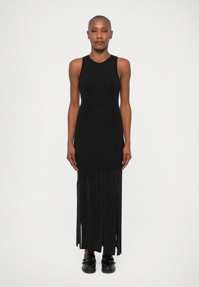 Robe sans manches noire avec un haut côtelé et un ourlet frangé asymétrique. Texture lisse, longueur cheville, agrémentée de chaussures noires avec des boucles.