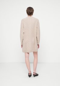 Rochie tricotată bej, cu mâneci lungi, având un model texturat și tiv ondulat, atingând lungimea de mijloc a coapsei.