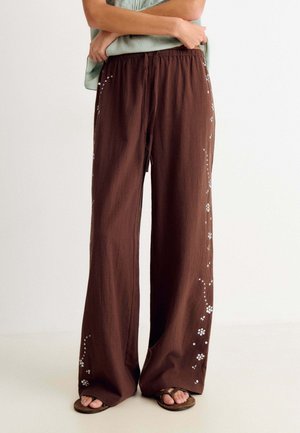 Persona con pantalones sueltos marrones con estampados florales blancos y sandalias, de pie con los brazos cruzados, sobre un fondo claro y liso.