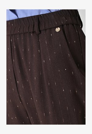 Gros plan sur un pantalon marron foncé à fines rayures discrètes, avec un petit bouton métallique rond près de la ceinture, associé à une chemise bleu clair.