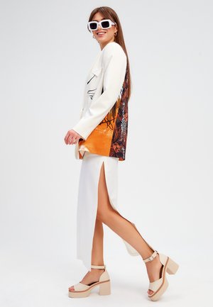 Witte blazer met kleurrijke, gemusterde achterkant, gedragen met een witte rok met zijsplitten. Beige plateausandalen met enkelbandjes.