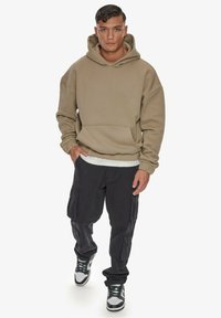 Hanorac oversized, de culoare maro deschis, cu buzunar frontal, asociat cu pantaloni cargo negri, ce au mai multe buzunare, și adidași gri și alb.