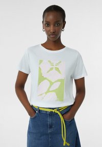 T-shirt en coton blanc avec un imprimé graphique vert et rose portant les mots « vacances faites à ». Associé à une jupe en denim et une ceinture jaune.