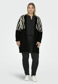 Zwart gebreid vest met een zebra stripe patroon in wit, open voorkant en geribde manchetten. Draagt met een zwarte top en zwarte jeans.