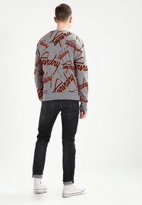 Uomo che indossa un maglione grigio a girocollo con testo arancione, jeans neri arrotolati e sneakers bianche alte, in piedi di spalle.
