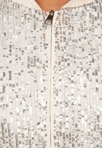 Casaco com lantejoulas, base em tecido creme com lantejoulas circulares prateadas e em rosa dourado, e fecho de correr frontal ao centro.