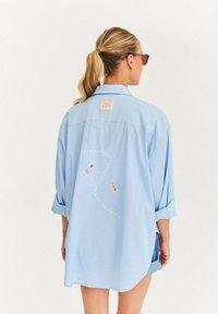 Žena s blond gumou na vlasy, ktorá má na sebe slnečné okuliare a svetlomodré voľné tričko s motívom plavcov a zakriveným textom na zadnej strane.