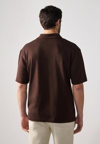 Polo shirt marron à manches courtes avec une texture douce, col classique et coupe décontractée. Porté avec un pantalon clair, montrant la vue arrière.