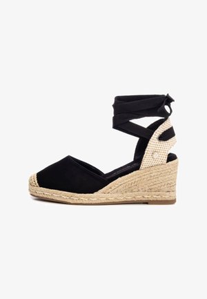 Zapato cuña espadrille negro con puntera redonda, suela de yute trenzado y tiras de tela cruzadas en el tobillo. Acento texturizado en el talón.