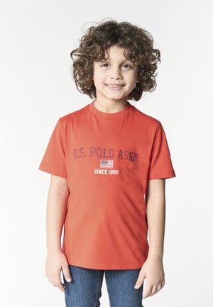 Bambino sorridente con capelli ricci, che indossa una maglietta arancione U.S. Polo Assn. e jeans blu, in piedi davanti a uno sfondo semplice.
