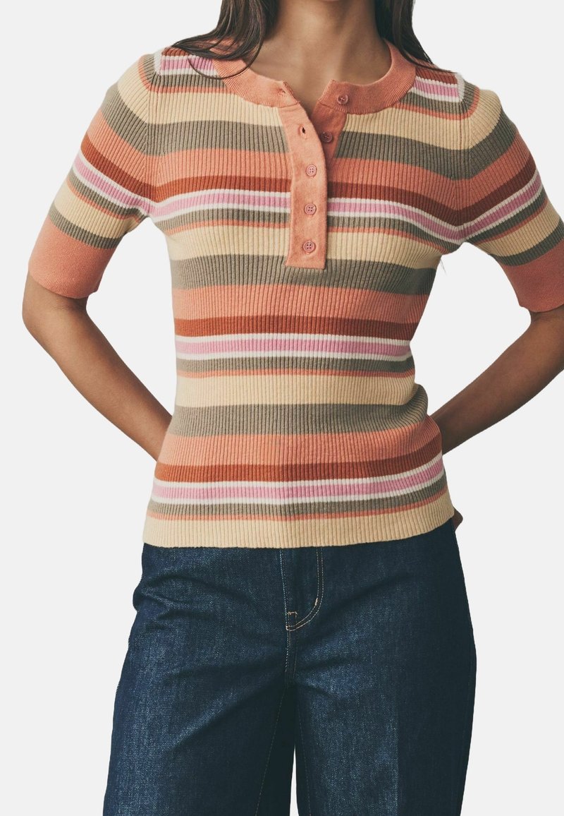 Femme portant un haut rayé à manches courtes côtelé aux tons orange, beige, rose et olive, associé à un jean en denim foncé, les mains sur les hanches.