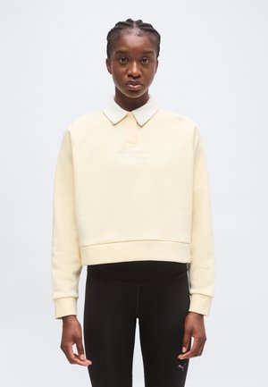 Ung kvinde med fletninger iført en cremefarvet langærmet pullover og sorte leggings, stående mod en ensfarvet baggrund.