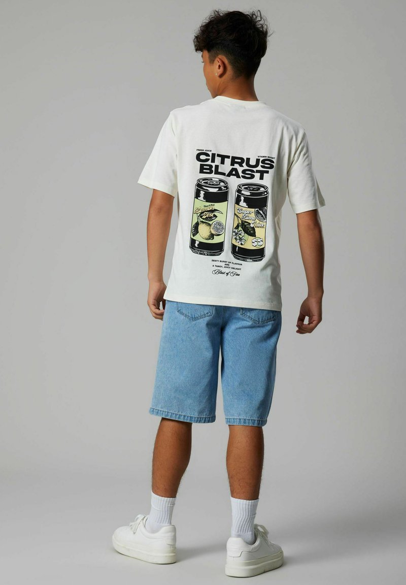 Personne portant un t-shirt blanc avec l'inscription "CITRUS BLAST" et un graphisme de canette de boisson aux agrumes dans le dos, un short en jean, des chaussettes blanches et des baskets blanches.
