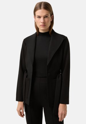 Sort blazer lavet af tekstureret stof med stor krave, enkelt knap og sidelommer. Kombineret med en sort rullekrave under.