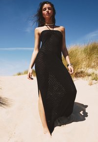 Robe maxi noire en crochet avec un design sans bretelles, des fentes sur les côtés et un motif texturé. Portée sur du sable dans un cadre extérieur naturel.