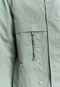 Gros plan sur la poche d'une veste vert clair avec une fermeture éclair verticale et un rabat boutonné, montrant la texture du tissu et les détails de la couture.