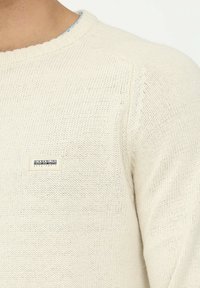 Cremfarbener gestrickter Pullover mit klassischem Rundhalsausschnitt, ausgestattet mit einem kleinen Markenpatch auf der linken Seite. Strukturiertes Material mit offenen Maschen.