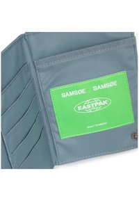 Portafoglio in tessuto grigio con molteplici fessure per carte, un'etichetta verde con il logo "EASTPAK" e il testo "SAMMØE". Materiale strutturato e chiusura a scatto.