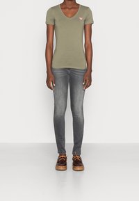 Olive green V-neck t-shirt, slim-fit gray jeans, and brown shoes with a tan sole. Notable t-shirt logo detail in red.