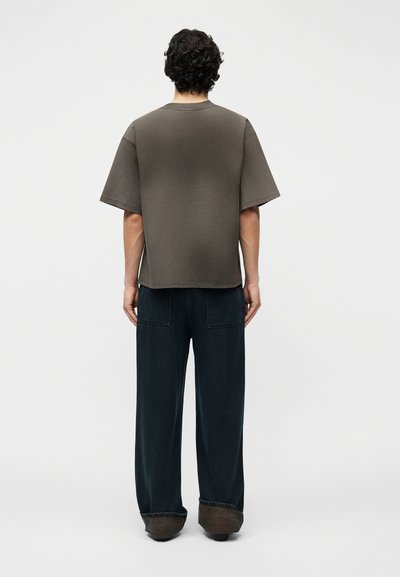 Άνδρας με σγουρά μαλλιά που στέκεται με την πλάτη, φοράει χαλαρό σκουρό γκρι μπλουζάκι και φαρδιά μαύρη παντελόνια με γυρισμένα μανίκια.