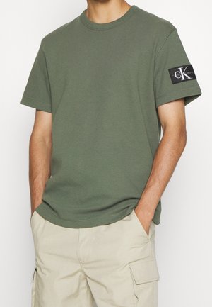 Mann trägt olivgrünes kurzarm T-Shirt mit Calvin Klein Jeans-Logo auf dem Ärmel und beige Cargo-Shorts, Hände in den Taschen.