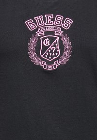 Camiseta de algodón negra con un logo bordado en rosa que presenta un diseño de escudo, hojas decorativas y el texto "GUESS LOS ÁNGELES 1981."