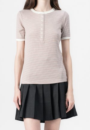 Femme portant un t-shirt henley à manches courtes rayé de couleur claire associé à une mini-jupe plissée noire sur fond blanc.