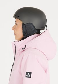 Black ski helmet with ventilation, worn with a light pink waterproof jacket featuring a drawstring hood and a logo patch on the sleeve.
