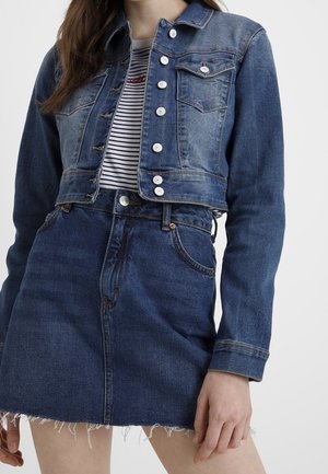 Kvinde iført en tofarvet cropped denimjakke, stribet skjorte og mørkeblå denim mininederdel med frynset kant mod en ensfarvet baggrund.