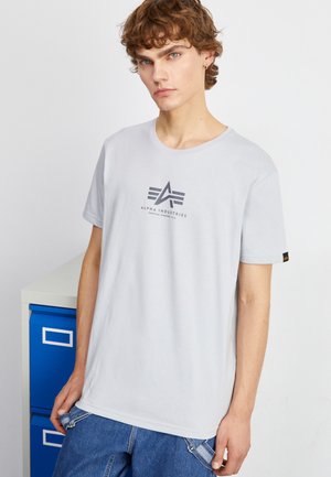 Helles graues T-Shirt mit Rundhalsausschnitt, das ein dunkles Logo und Text in der Mitte zeigt. Das Material wirkt weich; kombiniert mit blauen Denim-Hosen.