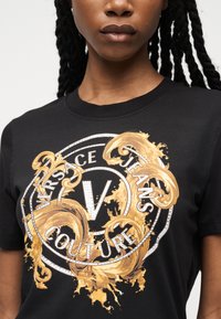 Μαύρο μπλουζάκι με χρυσούς μπαρόκ στροβίλους και λευκό κυκλικό λογότυπο Versace Jeans Couture στο στήθος, που φοριέται από ένα άτομο με μακριά κοτσίδα.