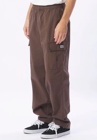 Bruna lösa cargobyxor med sidofickor, bärs av en person i svarta och vita sneakers och vit skjorta, står på vit bakgrund.