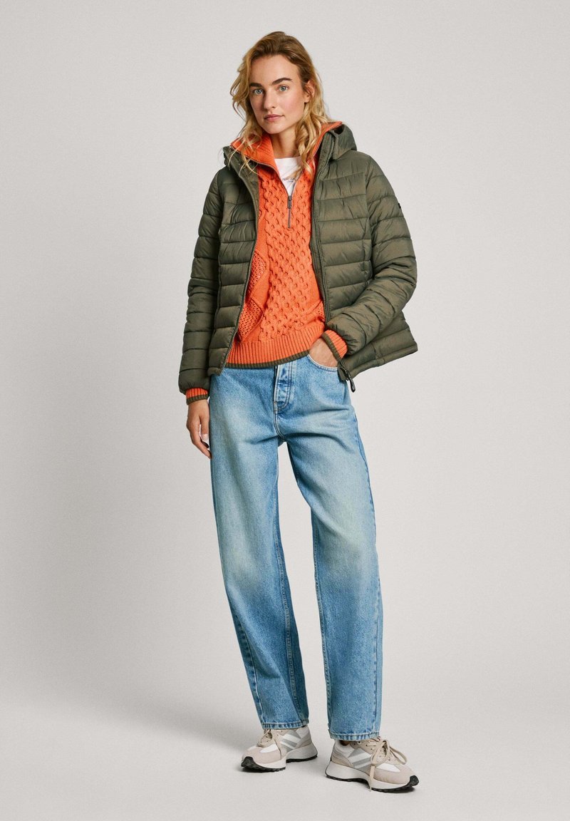 Doudoune vert olive avec capuche, superposée sur un pull en maille torsadée orange. Associée à un jean léger bleu clair et des baskets blanches.
