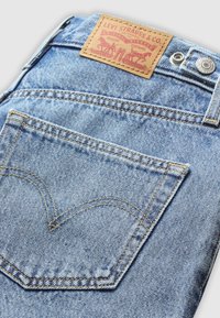 Vaaleansiniset farkut, joissa on ruskea nahkalamppu, jossa lukee "Levi Strauss & Co." ja keltaiset ompeleet. Takapussissa on vaikuttava design.