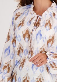 Langslibblouse van lichtgewicht stof, met een witte ondergrond en abstracte patronen in bruin en blauw, een fluffy halslijn en een strikdetail.