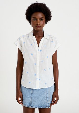 Vrouw staand tegen een witte achtergrond, gekleed in een witte blouse met korte mouwen en blauwe patronen en een lichtblauwe spijkerminirok.