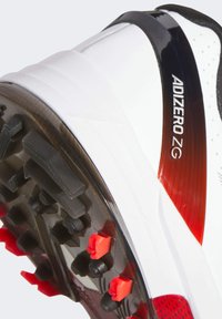Białe buty sportowe z teksturowanym wzorem, z gumową podeszwą, czarnymi akcentami, czerwonymi wstawkami oraz brandingiem "ADIZERO ZG" z boku.