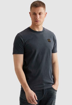 Jeune homme aux cheveux courts portant un t-shirt gris foncé à manches courtes et un jean foncé, regardant légèrement vers le bas et sur le côté.