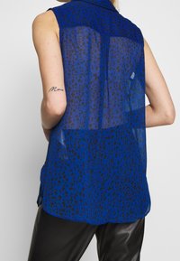 Top sin mangas azul transparente con estampado de leopardo negro. Presenta un cuello con solapas y un corte relajado, resaltando una textura semitransparente.