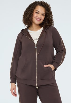 Donna dai capelli ricci che sorride, indossa una felpa marrone con zip e pantaloni coordinati con strisce laterali, posa con una mano in tasca.