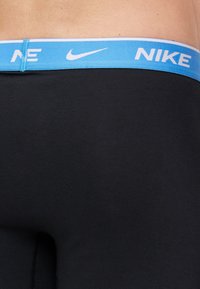 Juodos sportinės leggins algai su mėlynu elastingu juosmeniu, kuriame yra baltas "NIKE" logotipas ir balta juostelė. Lygi tekstūra, prigludusi konstrukcija.