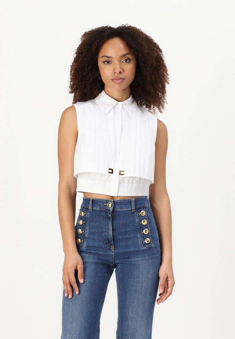 Camicia bianca cropped con colletto e pieghe verticali; abbinata a jeans blu a vita alta con dettagli di bottoni dorati.