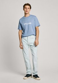Light blue cotton t-shirt with white "PEPE JEANS" print, paired with light wash denim jeans and dark sneakers with a cream sole.