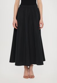 Svart maxi-skjørt laget av lett stoff, med høy elastisk midje, full silhuett og glatt tekstur. Brukt med beige sandaler.