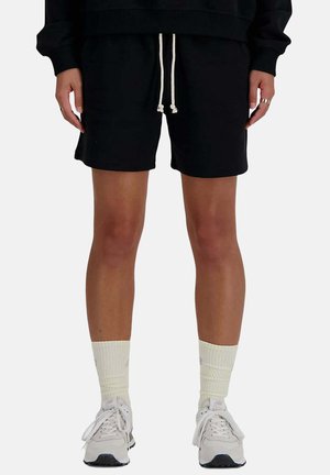 Persona che indossa pantaloncini neri con coulisse, calzini sportivi bianchi e sneakers bianche, in piedi con le mani rilassate lungo i fianchi.